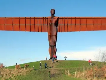 The Angel of the North sculpture stands on a green hill with people walking near its base under a clear blue sky.