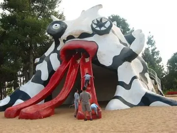 The Golem monster sculpture with three red slide-like tongues, with children climbing on it in a sandy park area.