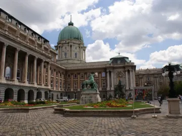 King Matthias Fountain sculpture group in a cobbled courtyard beside flowerbeds and a grand building with a green dome.