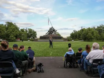 The Marine Corps War Memorial stands in a grassy park as a crowd sits facing a brass performer in the foreground.