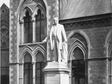 The Rolleston statue stands on a tall hexagonal pedestal behind a dark iron fence, with a stone building in the background.