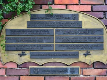 Commemorative plaque on the Warsaw Uprising Monument with Polish text and a small airplane relief, partly covered by ivy.