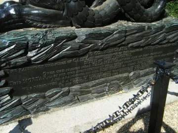 Close-up of the Cádiz Memorial plaque set into a sculpted base with textured relief and a dark metal post beside it.