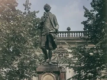 Goethe Monument on a tall granite pedestal with a golden plaque, surrounded by trees and a grand building in the background.