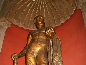 Hercules statue holding a club, with a lion skin and apples, set against a red wall beneath a gilded alcove.