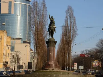 The Monument to Mykola Shchors, a bronze equestrian statue on a tall stone pedestal along a city street with trees and buildings nearby.