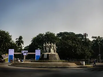 Raju Memorial Sculpture stands on a circular tiered base at a road junction, surrounded by trees, banners, and people nearby.