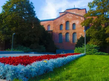 Unknown Soldier Monument beside a reddish-brick building with arched windows, framed by trees and flower beds.