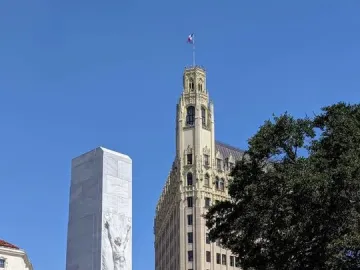 The Alamo Cenotaph stands in front of the Morgan Hotel tower under a clear blue sky.