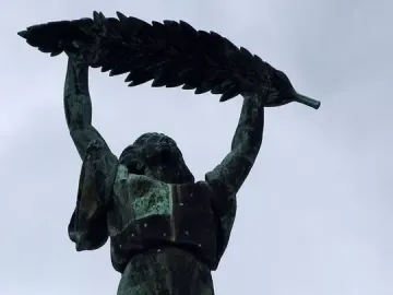 Liberty Statue bronze figure raising a leafy branch against a cloudy sky.