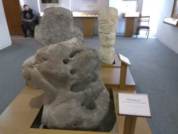 Škloŭ Idol stone sculpture with a rough grey surface and rounded indentations, displayed on a wooden stand.