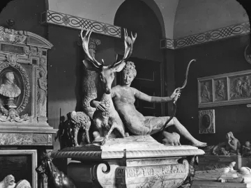 Black-and-white photograph of Diana with a stag sculpture, showing a reclining female figure beside a deer with large antlers in an interior gallery.