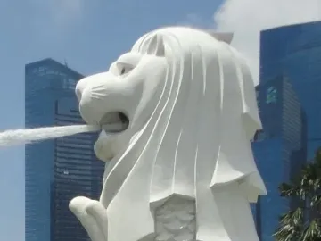 The Merlion statue with water jetting from its mouth, set against modern city buildings and a blue sky.