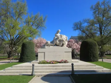 White stone war memorial with sculptural figures on a tiered base, surrounded by lawns and flowering trees.