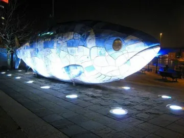The Big Fish sculpture covered in blue and white mosaic tiles, lit up at night on a waterfront plaza.
