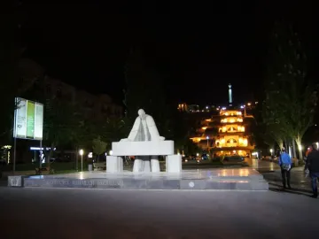 Monument to Alexander Tamanyan in a lit plaza at night, with a grand illuminated building complex and distant spire behind it.
