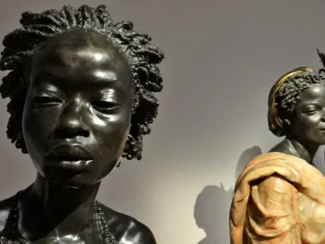The Nubian Woman bust with braided hair, closed eyes, and a beaded necklace, shown in front of two smaller sculptural busts.