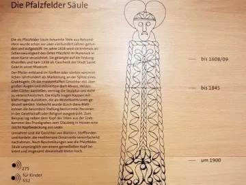Illustrated Pfalzfeld Obelisk shown in black outline with decorative faces and patterns on a light wooden background.