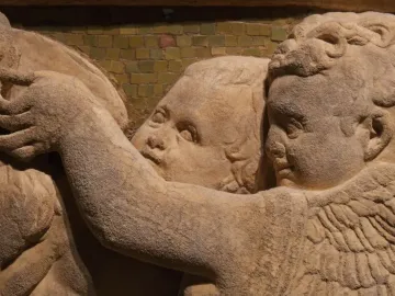 Close-up of a marble relief from the Pulpit of Prato Cathedral showing winged figures, faces, and a hand in front of a mosaic background.