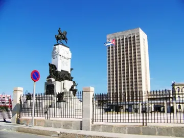 Antonio Maceo Monument with an equestrian statue on a tall white pedestal, a high-rise building and Cuban flag behind it.