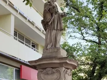 Weathered stone statue of John of Nepomuk on a sculpted pedestal behind a decorative metal fence.