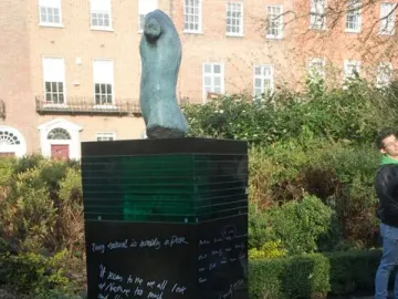Oscar Wilde Memorial Sculpture on a tall dark pillar with white inscriptions beside a paved path and greenery.