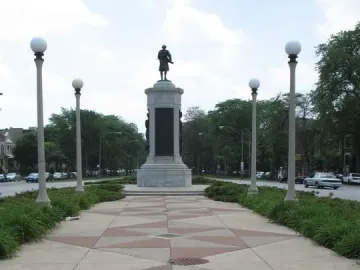 Victory Monument rising at the end of a patterned pedestrian walkway with lampposts and trees nearby.