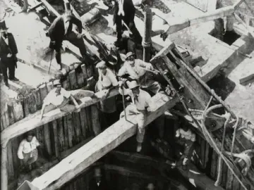 A large reclining Hercules statue is being unearthed from a timber-lined excavation pit as several men work around it.