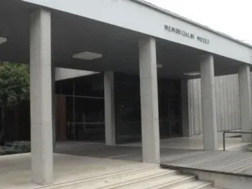 Front entrance of the Memorial Museum Jasenovac with grey concrete pillars, glass windows, and steps leading up to the door.