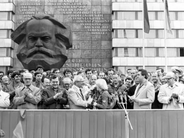 Karl Marx Monument beside a crowd gathered for an outdoor event, with a building facade behind it bearing a multilingual slogan.
