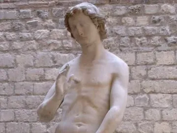 Stone statue of Adam standing nude with a leaf covering his groin and one hand raised.