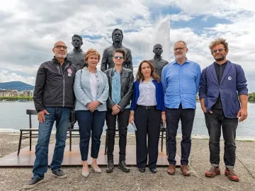 Seven people pose beside the Anything to Say? sculpture with three seated bronze figures and an empty chair by the water in Geneva.