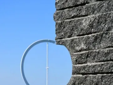 The Éole ring sculpture is seen through an opening in a dark stone wall, with water and sky behind it.