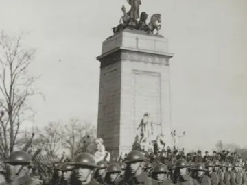 USS Maine National Monument with a sculptural figure and horses atop a tall stone base, seen behind marching soldiers.