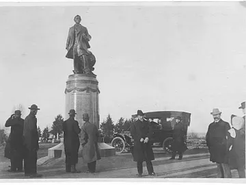 William Henry Seward monument in Volunteer Park with several men gathered around the base and a vintage automobile nearby.