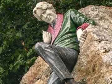 Oscar Wilde Memorial Sculpture showing a reclining man beside a rough boulder with leafy trees behind him.