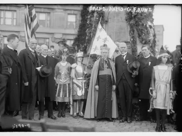A dark Joan of Arc monument draped with garlands and foliage stands behind a cardinal and children in historical costumes.
