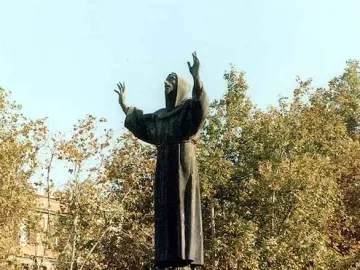 Monument to Saint Francis of Assisi, a dark bronze hooded figure with raised hands against a clear sky and autumn trees.