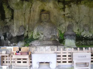 Usuki Stone Buddhas carved into a rock face, with a central seated figure and smaller figures nearby beside offerings and a wooden fence
