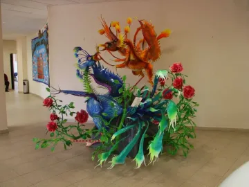 Alebrije sculpture with a blue-purple winged creature beside an orange-red bird-like figure, surrounded by green foliage and red roses.