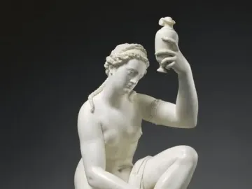 White sculpture of a kneeling female figure holding a small vessel aloft beside a draped pillar.