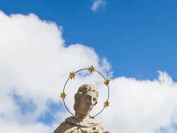 Statue of St. John of Nepomuk viewed from below against a blue sky, wearing a halo of stars and holding an object to his chest.