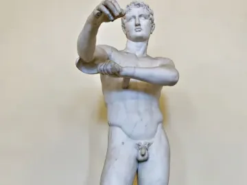 White marble Apoxyomenos statue of a nude male figure standing with one leg bearing his weight and his arms raised.