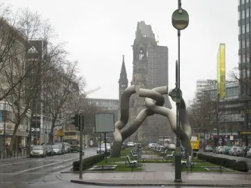 Light-grey tubular sculpture by Brigitte und Martin Matschinsky-Denninghoff in a city plaza, with a historic church ruin in the background.