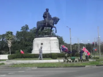 Gen. Beauregard equestrian statue on a tall white pedestal in a grassy traffic circle, with several people nearby displaying Confederate battle flags.
