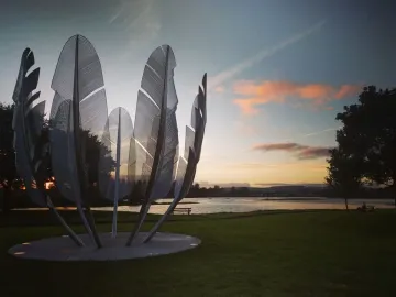Kindred Spirits sculpture with curved metallic feather-like forms beside the water at sunset.