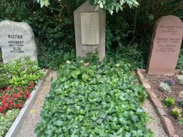 Mourning Athena stone relief on a vertical headstone beside a small plaque and greenery.