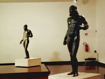 Two Riace bronzes on pedestals in a gallery, with one larger bearded nude male statue in the foreground and a smaller figure behind it.