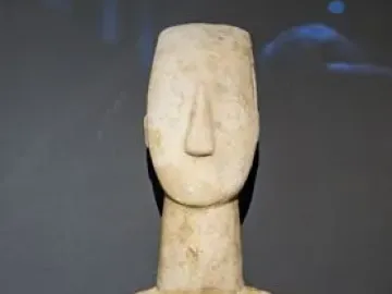 Kykladic idol with folded arms and a long, narrow marble body against a dark background.