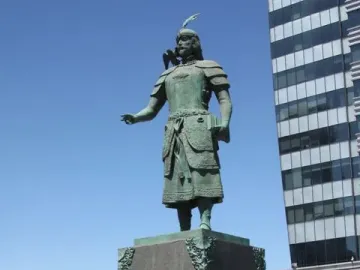 Marco Polo statue on a stone pedestal, with a bronze figure holding a book under a blue sky.